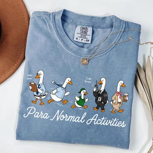 Puede incluir: Una camiseta azul con un gráfico de cinco patos de dibujos animados vestidos con varios atuendos y el texto "Para Normal Activities" impreso debajo. El primer pato sostiene un portapapeles, el segundo lleva un cordón, el tercero una gorra verde, el cuarto un walkie-talkie y el quinto una taza de café. La camiseta tiene una etiqueta de la marca Comfort Colors.