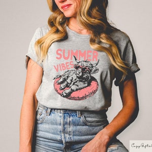 May include: A gray t-shirt with a pink graphic of a highland cow wearing sunglasses and floating in a pink inner tube. The text "Summer Vibes" is printed in pink above the cow.