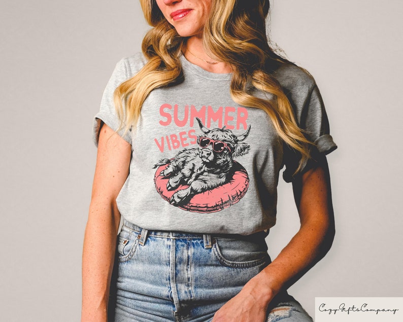May include: A gray t-shirt with a pink graphic of a highland cow wearing sunglasses and floating in a pink inner tube. The text "Summer Vibes" is printed in pink above the cow.