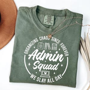 Könnte beinhalten: Ein olivgrünes T-Shirt mit dem Text "ORGANIZING CHAOS SINCE FOREVER Admin Squad WE SLAY ALL DAY" in einem weißen kreisförmigen Design. Eine goldene Halskette und ein brauner Hut sind im Hintergrund zu sehen.