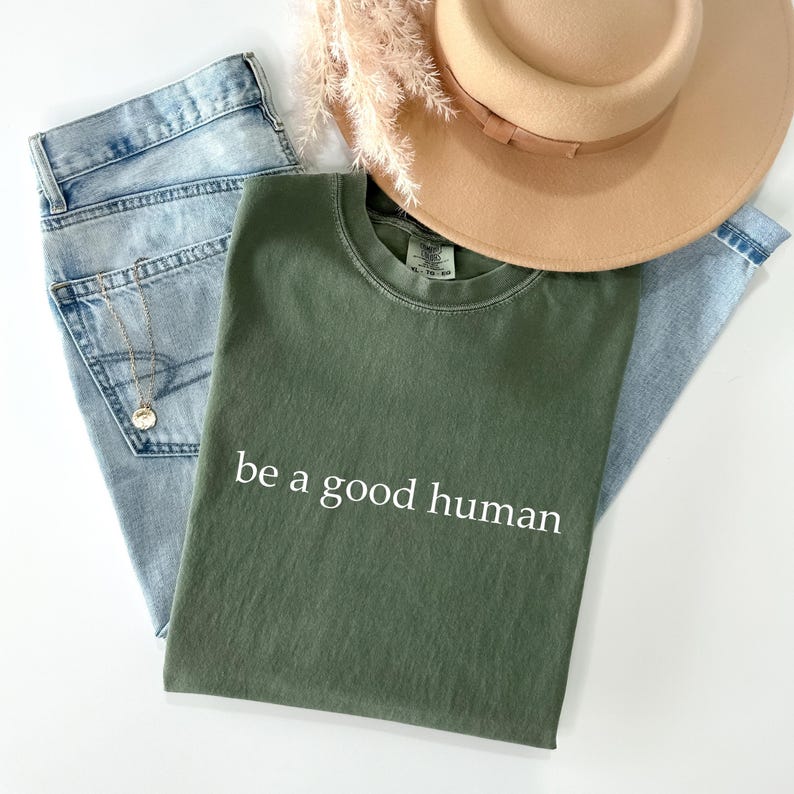 May include: An olive green t-shirt with the text "be a good human" in white, paired with light wash denim jeans, a gold necklace, and a tan hat. Dried flowers add a touch of natural decor.
