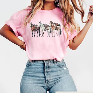 May include: A pink t-shirt with a graphic of four horses wearing saddles. The horses are brown, white, and black. The t-shirt is being worn by a person with long brown hair.