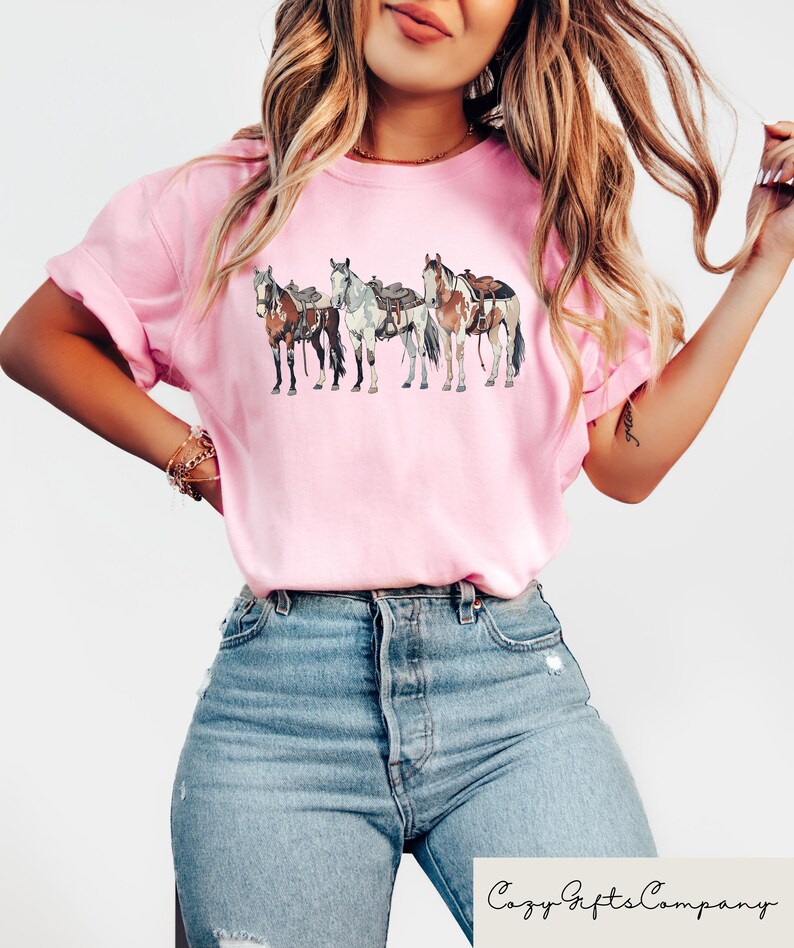 May include: A pink t-shirt with a graphic of four horses wearing saddles. The horses are brown, white, and black. The t-shirt is being worn by a person with long brown hair.