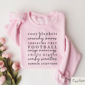 Op de afbeelding: Roze sweatshirt met de tekst "cozy blankets crunchy leaves crackling fires football crisp mornings chilly nights comfy sweaters pumpkin everything" gedrukt op de voorkant.