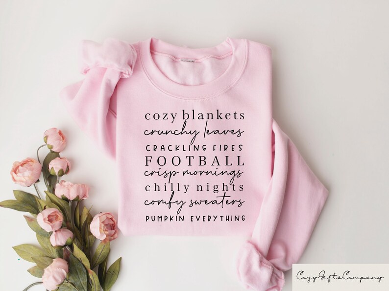 Op de afbeelding: Roze sweatshirt met de tekst "cozy blankets crunchy leaves crackling fires football crisp mornings chilly nights comfy sweaters pumpkin everything" gedrukt op de voorkant.