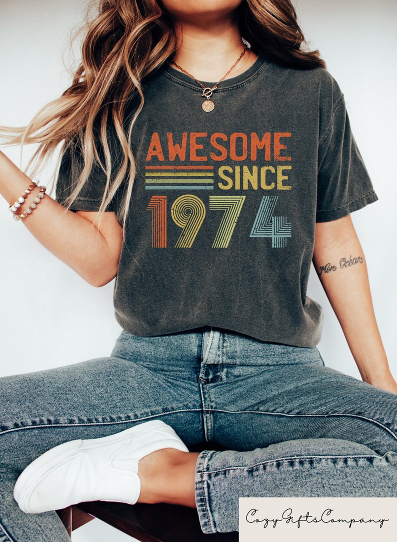 Pu&ograve; includere: Una maglietta grigia scura con il testo "Awesome Since 1974" in un design arcobaleno retr&ograve;.