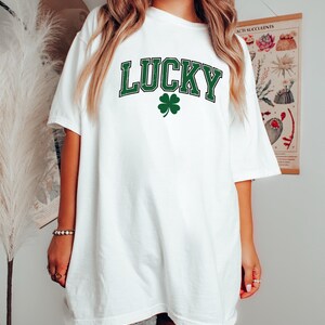 K&ouml;nnte beinhalten: Wei&szlig;es T-Shirt mit dem Wort "LUCKY" in gr&uuml;ner Schrift und einem gr&uuml;nen Kleeblatt. Das Shirt hat eine lockere Passform, ideal f&uuml;r den Alltag oder f&uuml;r St. Patrick's Day.
