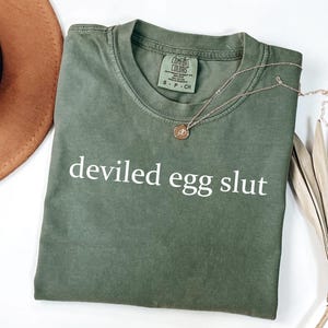 Puede incluir: Camiseta verde oliva con la frase "deviled egg slut" en letras blancas. Un collar dorado con un colgante descansa sobre la camiseta. Un sombrero marr&oacute;n y una planta seca est&aacute;n en el fondo. La camiseta est&aacute; doblada.