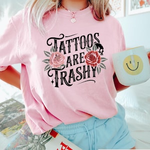 May include: Pink t-shirt with a black graphic that says "Tattoos are trashy" with floral accents.