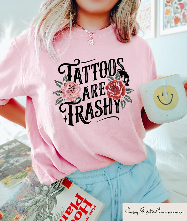 May include: Pink t-shirt with a black graphic that says "Tattoos are trashy" with floral accents.