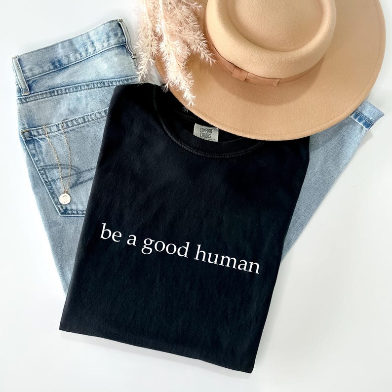 May include: A black t-shirt with the phrase "be a good human" in white lettering. The shirt is laid on top of light blue jeans, with a tan hat and a sprig of dried flowers. A gold necklace is visible.