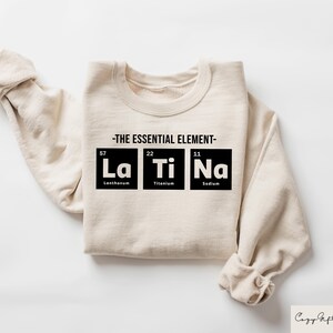 May include: A beige sweatshirt with the text "THE ESSENTIAL ELEMENT" and the chemical symbols for Lanthanum, Titanium, and Sodium, with their atomic numbers and names below.