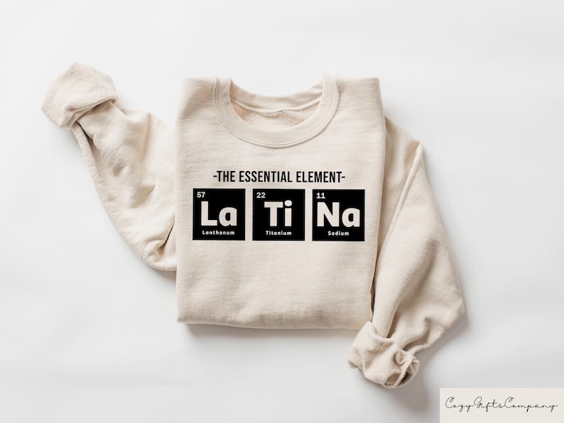 May include: A beige sweatshirt with the text "THE ESSENTIAL ELEMENT" and the chemical symbols for Lanthanum, Titanium, and Sodium, with their atomic numbers and names below.