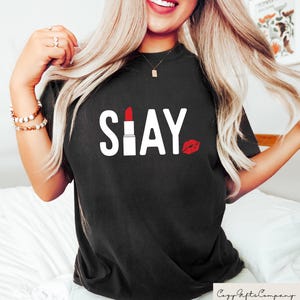 Puede incluir: Camiseta negra con la palabra "SLAY" en blanco. La "I" de "SLAY" es un l&aacute;piz labial rojo, y una marca de beso de l&aacute;piz labial rojo est&aacute; al lado de la palabra. La camiseta es de estilo casual.