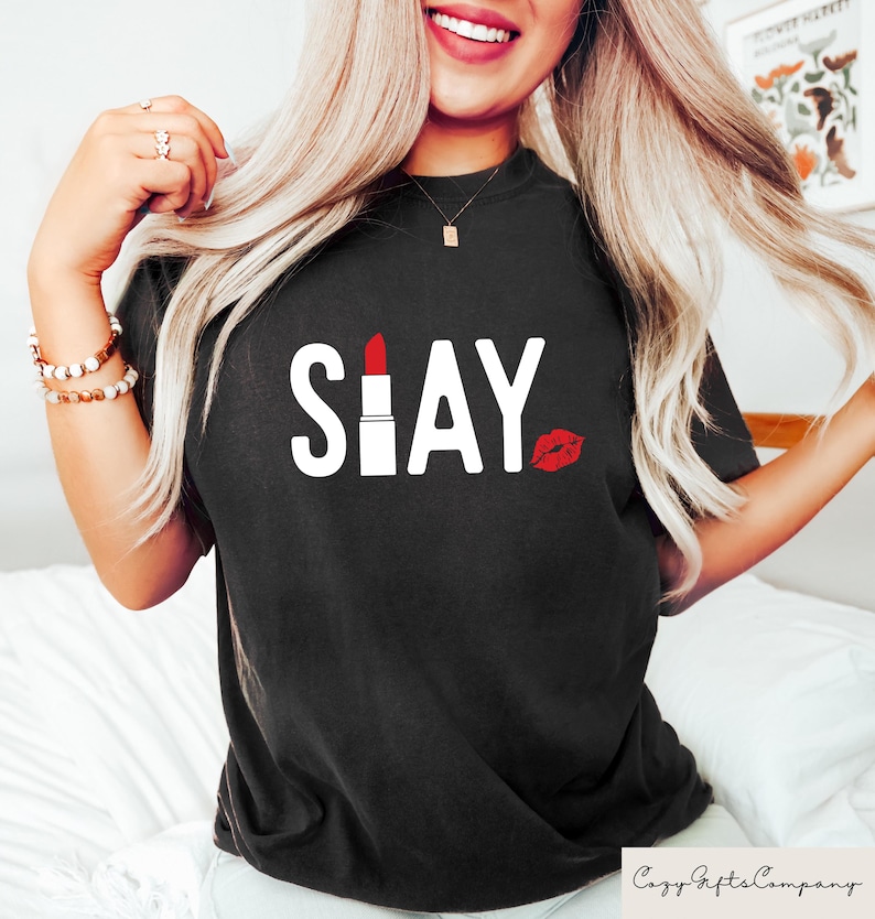 Puede incluir: Camiseta negra con la palabra "SLAY" en blanco. La "I" de "SLAY" es un l&aacute;piz labial rojo, y una marca de beso de l&aacute;piz labial rojo est&aacute; al lado de la palabra. La camiseta es de estilo casual.