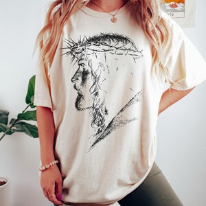 May include: Cream-colored t-shirt featuring a black and white illustration of a man wearing a crown of thorns. The graphic is a side profile of the man's face. The shirt has short sleeves and a relaxed fit.
