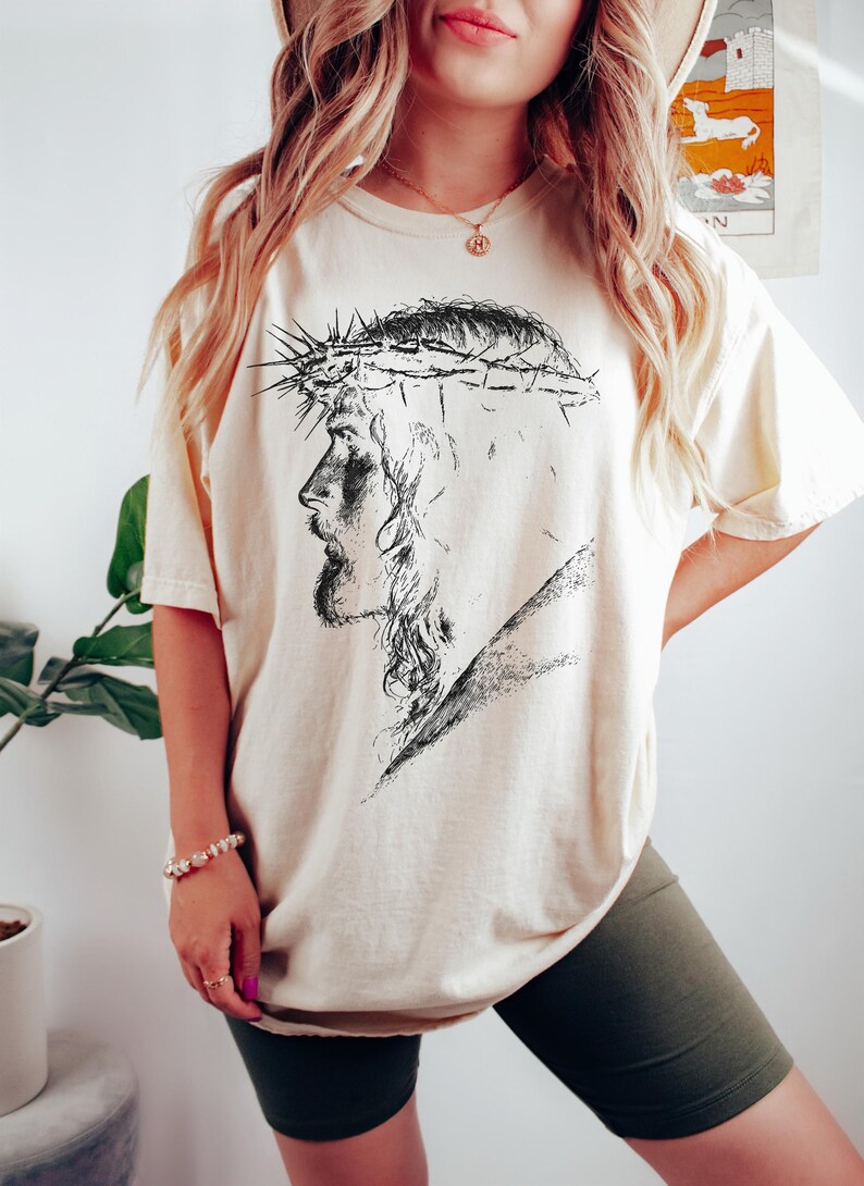 May include: Cream-colored t-shirt featuring a black and white illustration of a man wearing a crown of thorns. The graphic is a side profile of the man's face. The shirt has short sleeves and a relaxed fit.