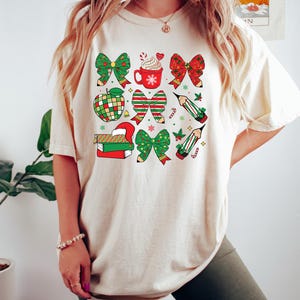 Puede incluir: Una camiseta de color crema con un dise&ntilde;o navide&ntilde;o con lazos rojos y verdes, l&aacute;pices, una taza de chocolate caliente, una manzana y libros.