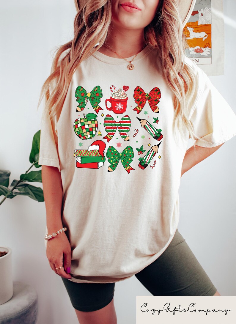 Puede incluir: Una camiseta de color crema con un dise&ntilde;o navide&ntilde;o con lazos rojos y verdes, l&aacute;pices, una taza de chocolate caliente, una manzana y libros.