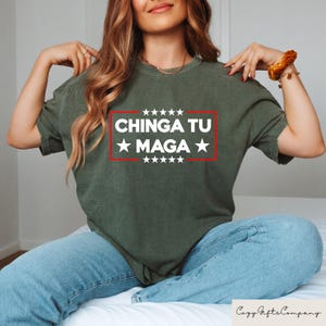 K&ouml;nnte beinhalten: Olivgr&uuml;nes T-Shirt mit dem Text &bdquo;CHINGA TU MAGA&ldquo; in wei&szlig;en Buchstaben in einem roten Rechteck, mit wei&szlig;en Sternen &uuml;ber und unter dem Text. Das Shirt wird mit einer blauen Jeans getragen.