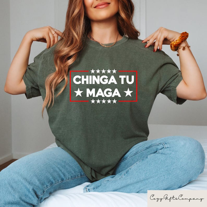 K&ouml;nnte beinhalten: Olivgr&uuml;nes T-Shirt mit dem Text &bdquo;CHINGA TU MAGA&ldquo; in wei&szlig;en Buchstaben in einem roten Rechteck, mit wei&szlig;en Sternen &uuml;ber und unter dem Text. Das Shirt wird mit einer blauen Jeans getragen.