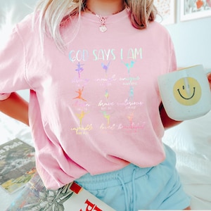 May include: A light pink t-shirt with the text "GOD SAYS I AM" and colorful ballet dancer illustrations. The shirt also features the words "amazing, novel, unique, loved, brave, victorious, capable, loved, beautiful". A white mug with a yellow smiley face is held in the hand.