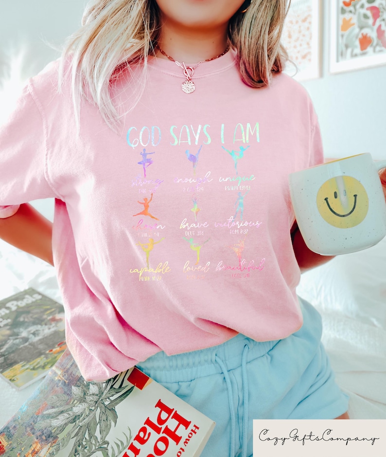 May include: A light pink t-shirt with the text "GOD SAYS I AM" and colorful ballet dancer illustrations. The shirt also features the words "amazing, novel, unique, loved, brave, victorious, capable, loved, beautiful". A white mug with a yellow smiley face is held in the hand.
