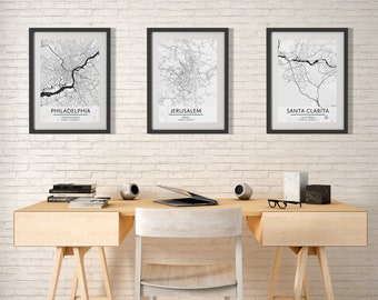 May include: A home office workspace with a white brick wall, a wooden desk with two drawers, a white chair, a yellow pendant light, and three framed black and white city maps with the names "Philadelphia", "Jerusalem", and "Santa Clarita" printed below the maps.