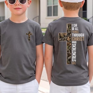 May include: A charcoal grey t-shirt featuring a small camouflage cross on the front left chest. The back displays a large camouflage cross with the white and tan text, I CAN DO ALL THINGS THROUGH CHRIST WHO STRENGTHENS ME, printed over the design.