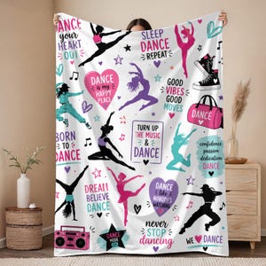May include: A white throw blanket featuring colorful dance-themed graphics, including silhouettes of dancers, hearts, stars, and musical notes. Text includes phrases like Dance Your Heart Out, Sleep Dance Repeat, and Dance Is My Happy Place.