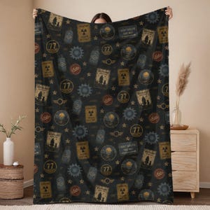 May include: A black throw blanket featuring a repeating pattern of Fallout-themed graphics including radiation hazard signs, the number 77, gear icons, and rectangular signs with the text Wasteland, Vault Shelter This Way, and Nuka.