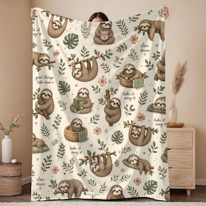 May include: A cream-colored throw blanket featuring a repeating pattern of brown sloths in various poses, including hanging from branches, sitting in baskets, and reading books, interspersed with green leaves, pink flowers, and the text take it easy.