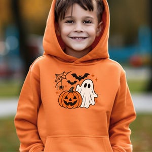 Puede incluir: Una sudadera con capucha de color naranja que presenta un estampado gráfico central de una calabaza sonriente, un fantasma blanco, murciélagos negros y una telaraña. La prenda incluye un bolsillo tipo canguro en la parte delantera.