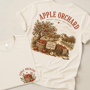 May include: A cream-colored t-shirt featuring a large back graphic of an apple orchard with a wooden sign reading PICK YOUR OWN HAPPINESS, a basket of red apples, and a cart. The front chest area displays a smaller graphic of an apple basket.