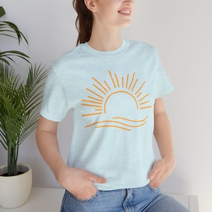 May include: Light blue t-shirt featuring an orange sun and wave design. The short-sleeved shirt is made of a soft material. The graphic is a simple line drawing of a sun setting over the ocean.