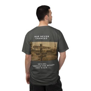 Könnte beinhalten: Dunkelgraues T-Shirt mit einem verblassten Bild eines Straßenschildes, das nach New Vegas, Commonwealth und Wasteland zeigt. Das Shirt trägt den Text "WAR NEVER CHANGES" über dem Bild und "MEN DO. THROUGH THE ROADS THEY WALK....." darunter.