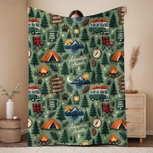 May include: A sage green fleece blanket featuring a repeating camping-themed pattern. The design includes green camper vans, orange tents, campfires, pine trees, lanterns, compasses, pinecones, and wooden signs with the text Adventure Awaits.