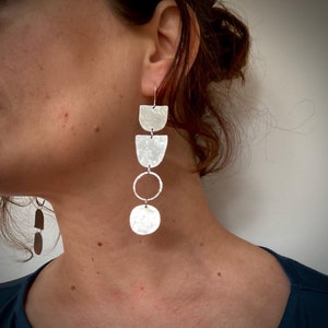 May include: Silver dangle earrings with a hammered finish. The earrings feature a series of three crescent shapes and a circle, followed by a larger round disc.