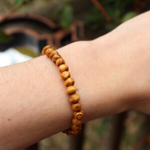 May include: A wooden bead bracelet with light brown, round beads. The bracelet is worn on a person's wrist, showcasing the natural wood grain and texture. The beads are strung together, creating a simple, earthy accessory.