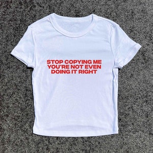 May include: White t-shirt with red text that reads "STOP COPYING ME YOU'RE NOT EVEN DOING IT RIGHT"