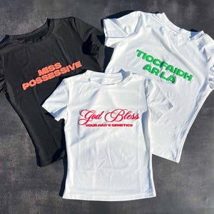 Może przedstawiać: Trzy białe koszulki z nadrukowanymi różnymi tekstami. Pierwsza koszulka ma napis "Miss Possessive" w różowych literach. Druga koszulka ma napis "God Bless Your Dad's Genetics" w czerwonych literach. Trzecia koszulka ma napis "Tiocfaidh ár lá" w zielonych literach.