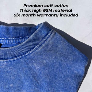 May include: Close-up of a blue t-shirt with the text "Premium soft cotton", "Thick high GSM material", and "Six month warranty included". The shirt has a textured appearance and a crew neck.