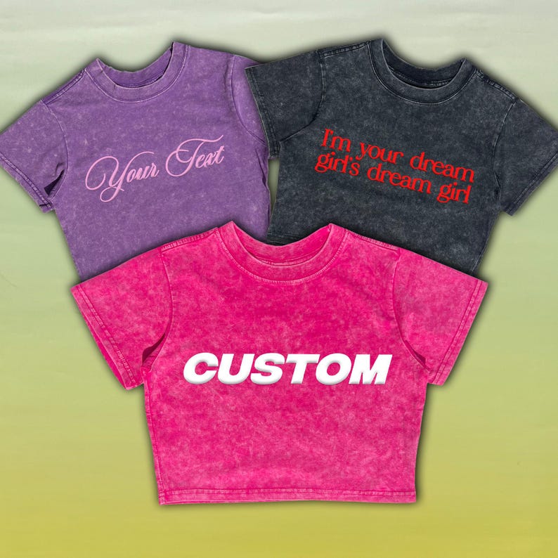 May include: Three short-sleeved t-shirts in purple, charcoal, and hot pink. The purple shirt has the text "Your Text" in a cursive font. The charcoal shirt has the text "I'm your dream girl's dream girl" in red. The hot pink shirt has the text "CUSTOM" in white.