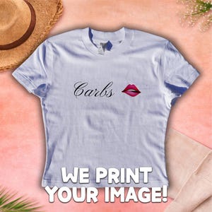 May include: A pale blue t-shirt featuring the word "Carbs" in cursive script and a red lipstick graphic. The shirt also displays the text "WE PRINT YOUR IMAGE!" in a bold, white font.