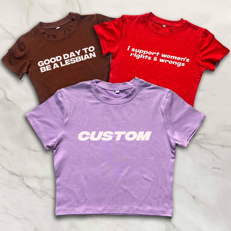 Puede incluir: Tres camisetas recortadas con diferentes lemas. La primera es marr&oacute;n con el texto "Good day to be a lesbian". La segunda es roja con el texto "I support women's rights & wrongs". La tercera es morada con el texto "CUSTOM" en blanco.