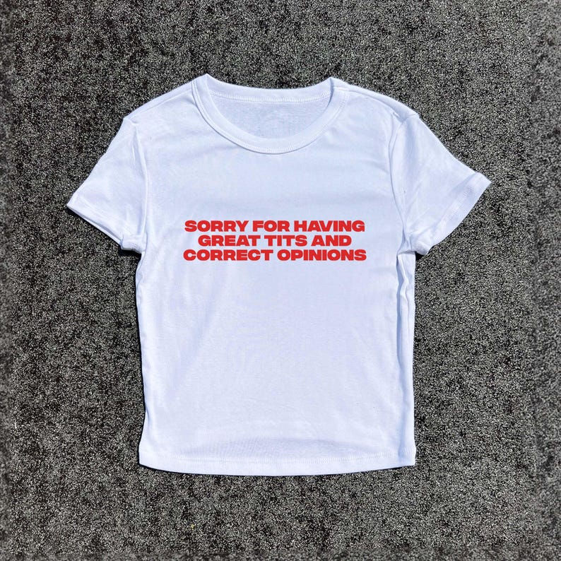 May include: White t-shirt with the text "SORRY FOR HAVING GREAT TITS AND CORRECT OPINIONS" in red. The shirt is made of a soft material and has short sleeves. The shirt is laying on a gray surface.