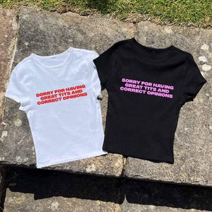 May include: Two t-shirts, one white and one black, with the text "SORRY FOR HAVING GREAT TITS AND CORRECT OPINIONS" printed on the front. The text on the white shirt is red, and the text on the black shirt is pink. Both shirts are short-sleeved.