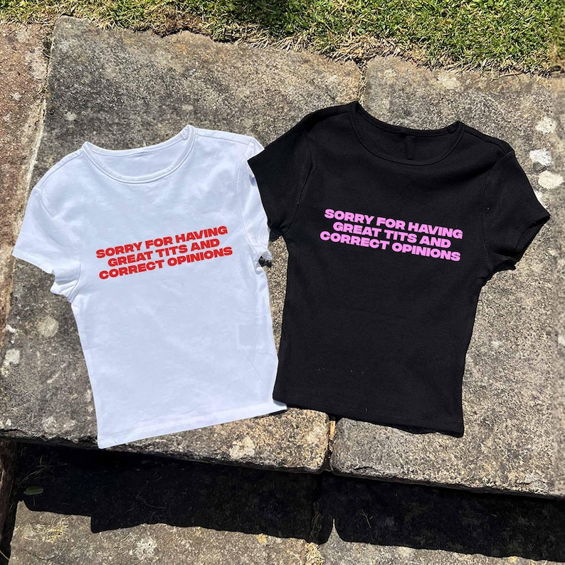 May include: Two t-shirts, one white and one black, with the text "SORRY FOR HAVING GREAT TITS AND CORRECT OPINIONS" printed on the front. The text on the white shirt is red, and the text on the black shirt is pink. Both shirts are short-sleeved.