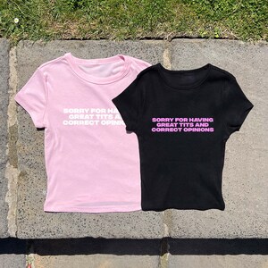 May include: Two t-shirts, one pink and one black, with the text "SORRY FOR HAVING GREAT TITS AND CORRECT OPINIONS" printed on the front. The pink shirt has white text, while the black shirt has pink text.