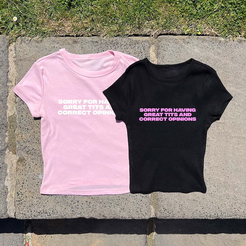 May include: Two t-shirts, one pink and one black, with the text "SORRY FOR HAVING GREAT TITS AND CORRECT OPINIONS" printed on the front. The pink shirt has white text, while the black shirt has pink text.